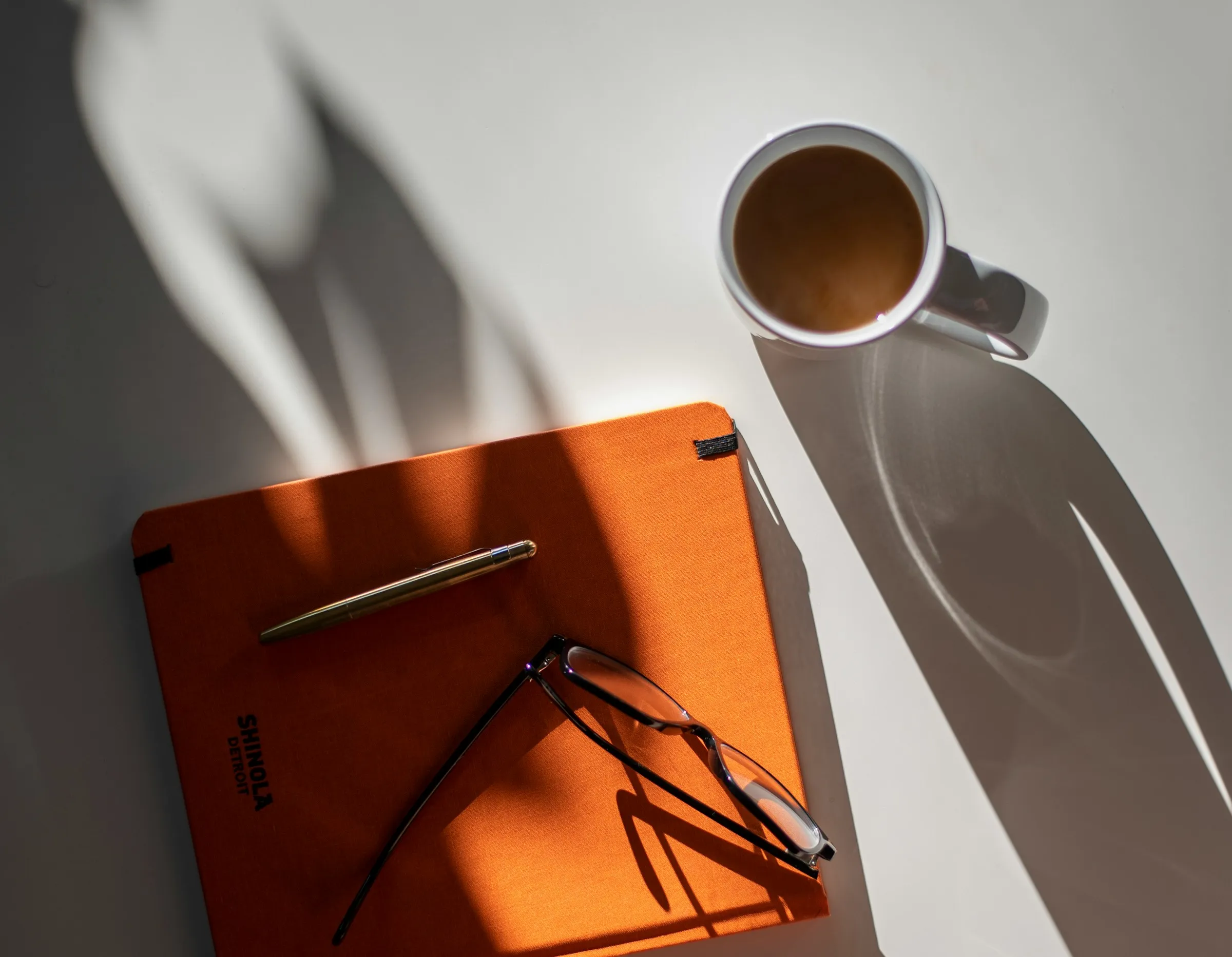A notebook, a pair of glasses and a coffee on a desk, with window shadows