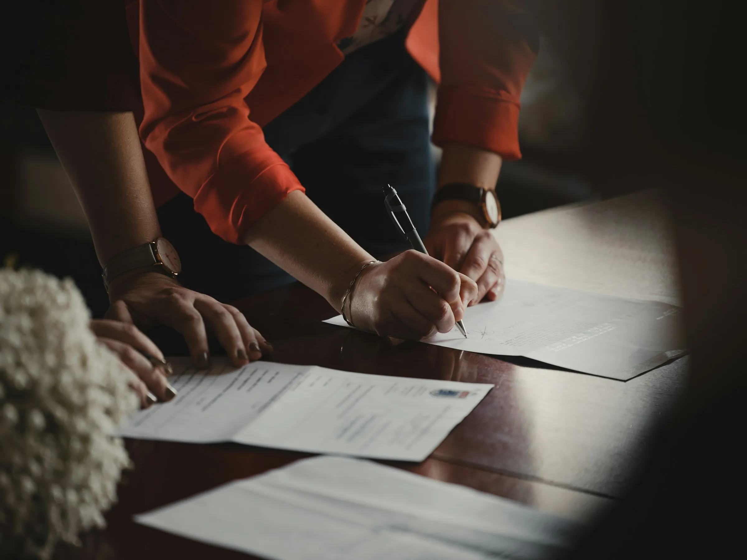 Dos personas firmando documentos sobre una mesa oscura, con luz cálida